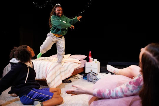 Three girls are in a bedroom; one stands and poses on the bed while the other two sit on the floor and a bean bag, with playful expressions. The room appears lively and casual.