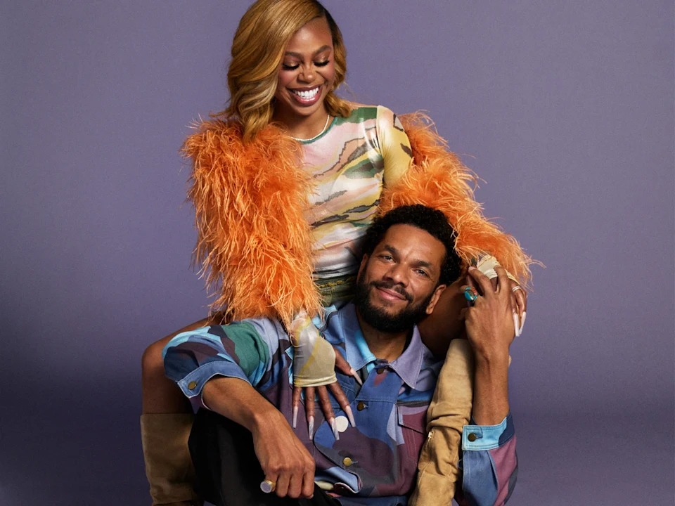 A woman in an orange feathered top sits behind a man in a patterned shirt, both smiling against a plain purple background.