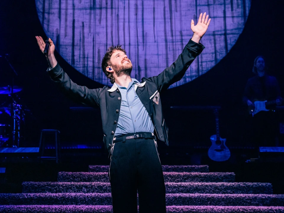 A man (Ben Platt) on stage with arms raised, wearing a blue shirt and black jacket, with a microphone headset. Background includes stairs, a large round light, a guitarist, and other musical equipment.