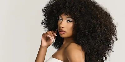 Woman with voluminous curly hair poses against a plain background, wearing a white off-shoulder top and natural makeup.