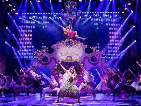 A dynamic stage performance features a cast of dancers in ornate costumes, with one performer suspended mid-air under a chandelier. The backdrop is elaborate with bright lighting effects.