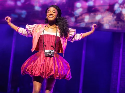 A person wearing a bright pink dress and jacket, with a camera hanging around their neck, poses with arms outstretched under colorful stage lighting.