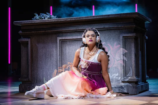 Production photo of & Juliet in New York, showing Gianna Harris sits on the floor in front of a large stone structure with pink and blue lighting in the background.
