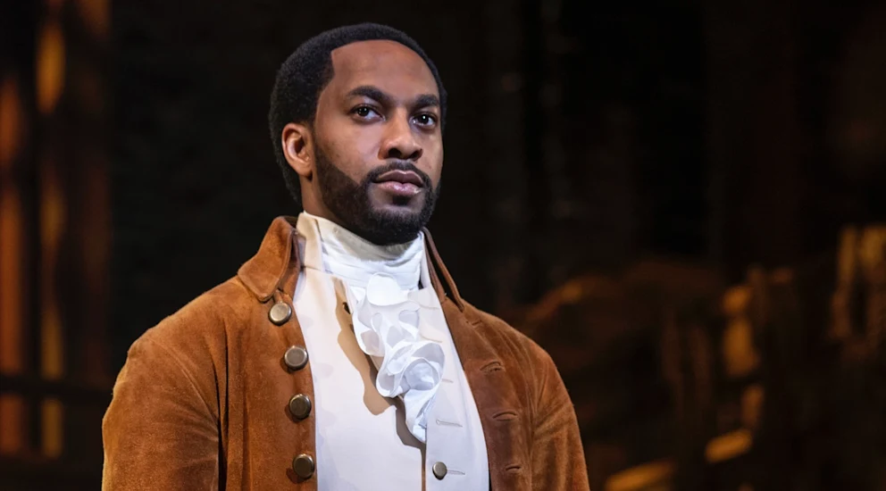A man dressed in historical 18th-century clothing with a brown coat, white vest, and cravat stands indoors, looking ahead with a serious expression.