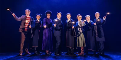 Eight people in costumes stand in a row on stage under blue lighting, each holding a lit wand and facing forward.