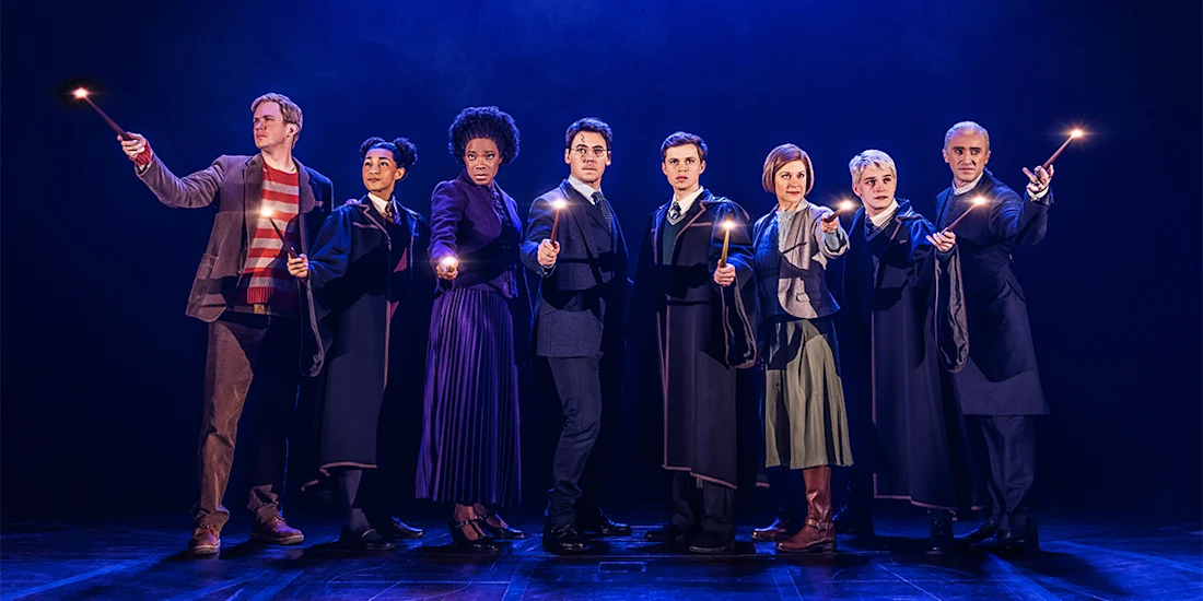 Eight people in costumes stand in a row on stage under blue lighting, each holding a lit wand and facing forward.