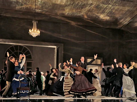 A group of people in period costumes dance energetically in a grand room with a chandelier and large windows.