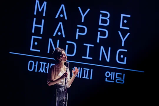 A woman stands at a microphone on stage in front of a sign that reads “Maybe Happy Ending” in English and Korean.
