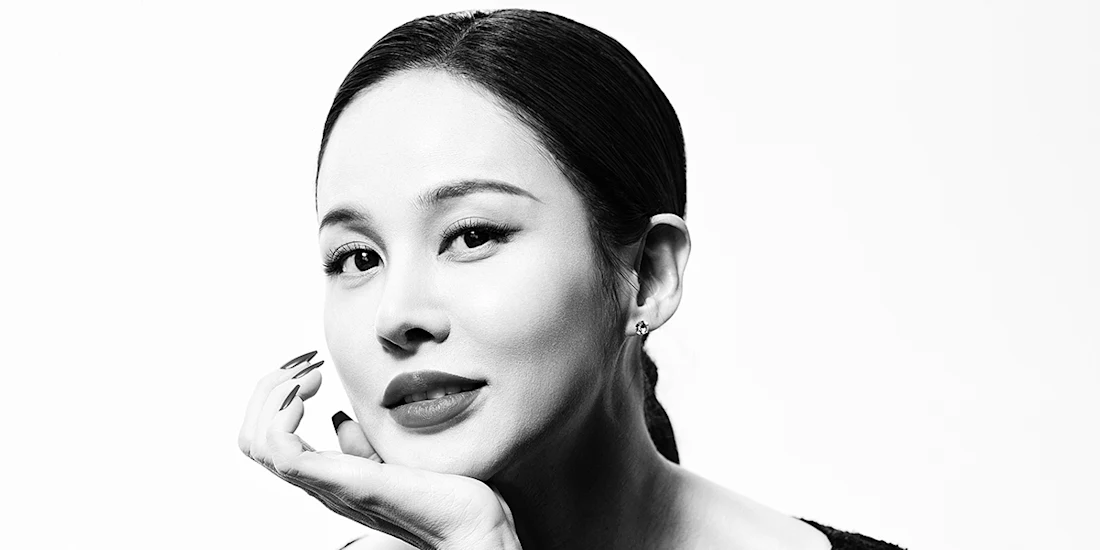 Black and white portrait of a woman with dark hair, wearing earrings, resting her chin on her hand, and looking at the camera with a slight smile.