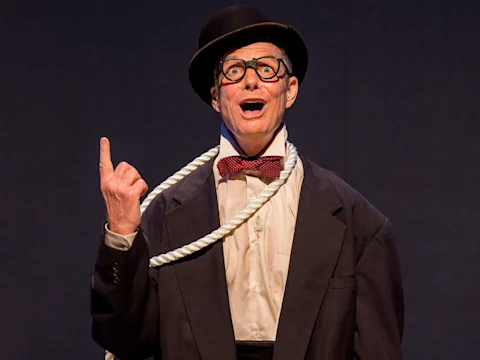 Man wearing a bowler hat, round glasses, and a suit with an oversized bow tie and rope around his neck, gesturing upward with his index finger against a dark background.