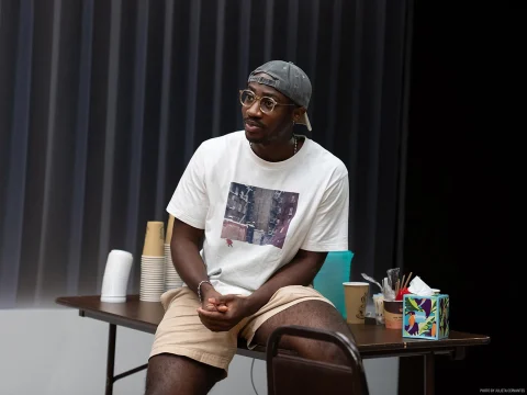 A person wearing a white t-shirt and shorts sits on a table with disposable cups and assorted items. They are also wearing glasses and a backwards baseball cap.