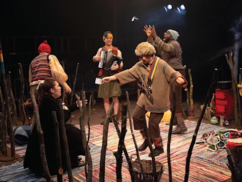Five people perform on a colorful stage with rugs and sticks; one plays an accordion, another juggles, and others interact with props under stage lighting.