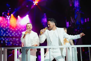 Two performers in white suits singing into microphones on stage under purple and red lights.