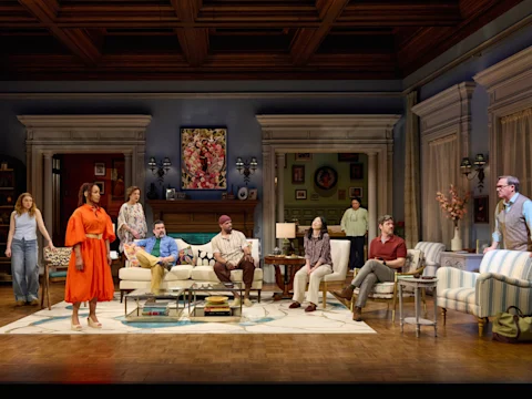 Eight people are gathered in a well-furnished living room, engaged in conversation, with some seated and others standing on a stage set for a theatrical performance.