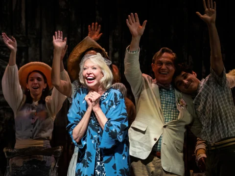 A group of actors on stage smiling and waving, with one woman in front clapping her hands, wearing a blue floral outfit.