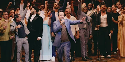 A man in a blue suit sings into a microphone on stage, surrounded by a group of cheering performers in theatrical costumes.