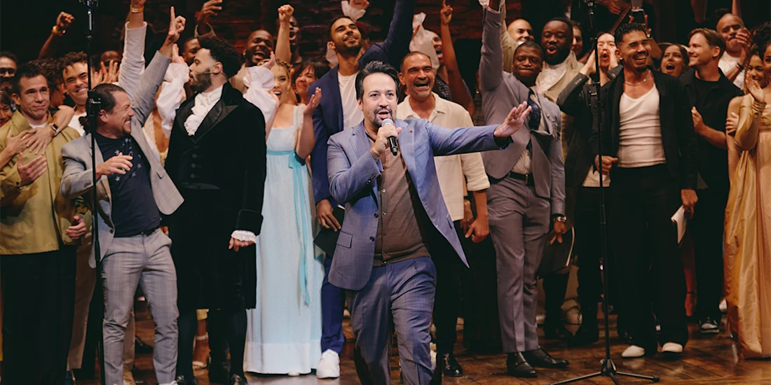 A man in a blue suit sings into a microphone on stage, surrounded by a group of cheering performers in theatrical costumes.