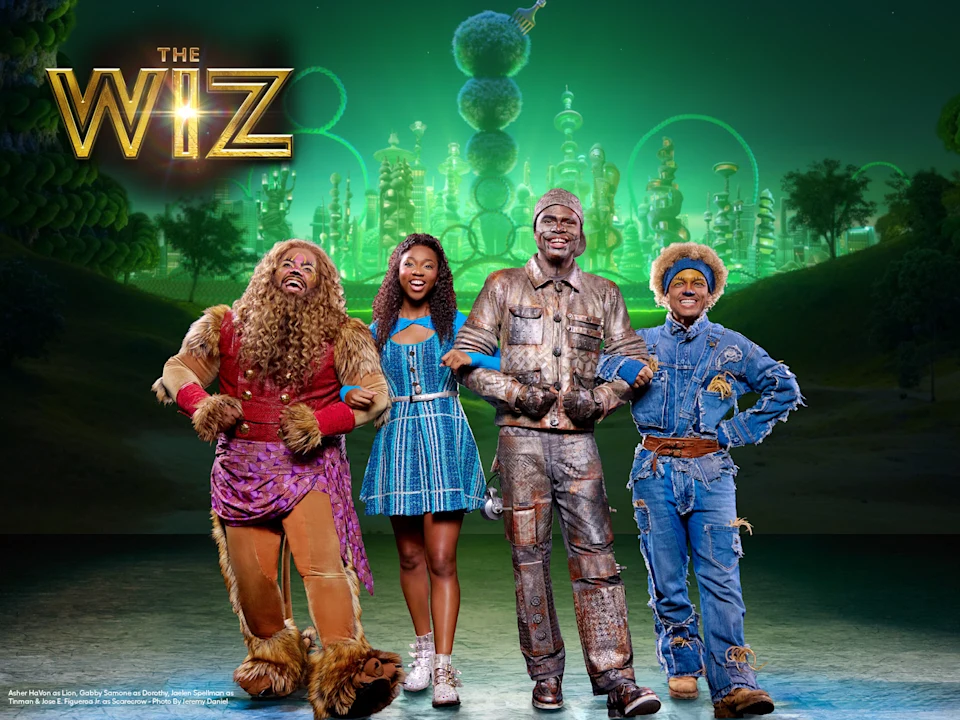 Four actors in colorful costumes pose smiling in front of a green, futuristic city backdrop under “The Wiz” logo.