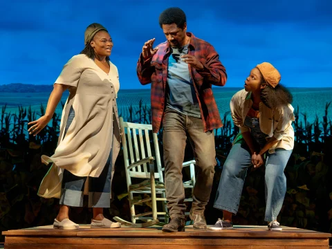 Three actors perform on stage against a backdrop of a cornfield, interacting with each other near a rocking chair.