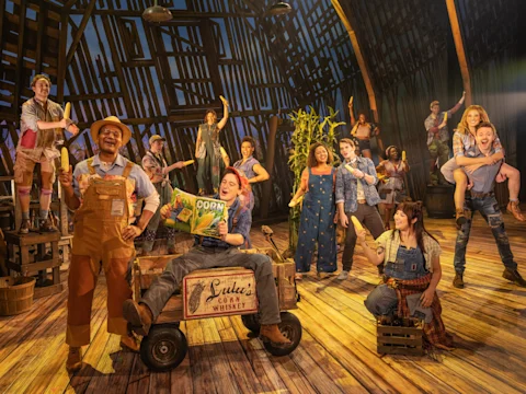 A group of actors in farm-themed costumes perform energetically on a wooden stage set resembling a barn, with props like corn, crates, and a wagon labeled "Lulu's Corn Whiskey.