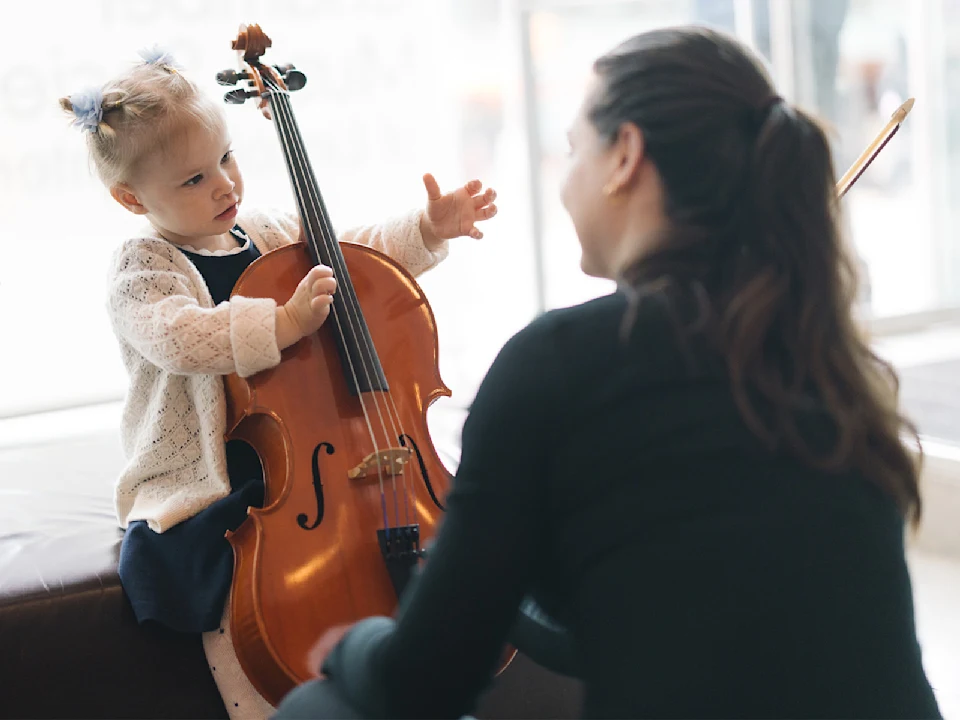 A young child sits holding a cello while an adult kneels in front of her, appearing to give guidance in a bright indoor setting.