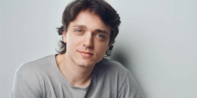 A young man with medium-length brown hair and light skin wearing a grey t-shirt looks directly at the camera against a plain light background.