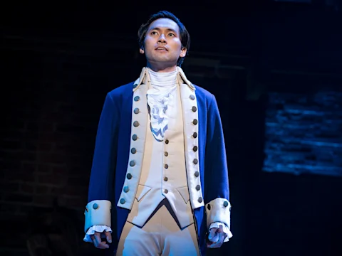 Production photo of Hamilton on Broadway, showing a person in a blue and cream historical costume stands on stage under dramatic lighting, with a dark background behind them.