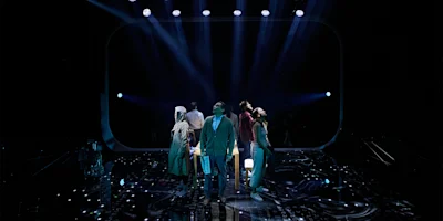 Six people stand in a circle under dramatic stage lighting, facing outward, on a dark stage with blue and purple lights overhead.