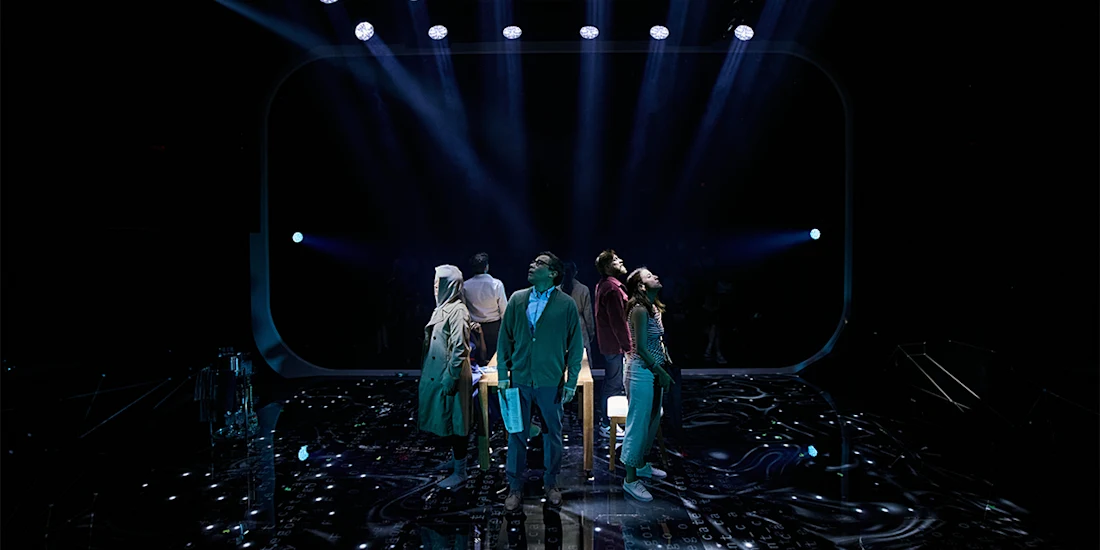 Six people stand in a circle under dramatic stage lighting, facing outward, on a dark stage with blue and purple lights overhead.