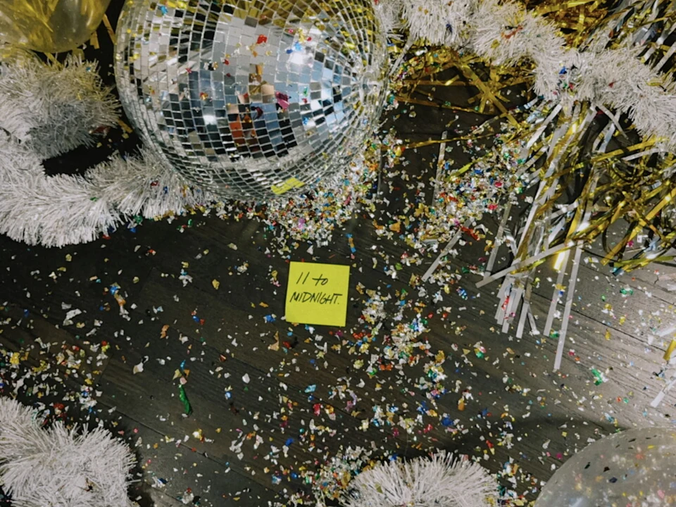 Disco balls, white garland, and gold streamers hang above a floor covered in confetti with a sticky note reading “11 to MIDNIGHT.”.