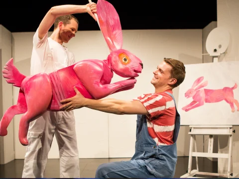 Two men hold a large, pink rabbit statue in front of a painting of the rabbit. One man wears a white outfit, and the other wears a red striped shirt and blue overalls.