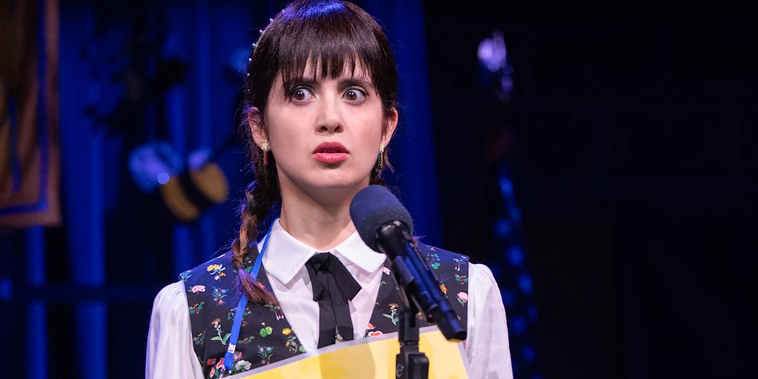 A young woman in a vest and necktie stands at a microphone, holding a folder and looking surprised on stage with dim blue lighting.
