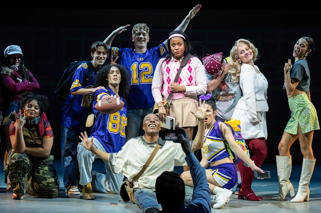 A group of actors in colorful costumes pose together on stage, some dressed as cheerleaders and football players, while a person in front takes a photo of them.