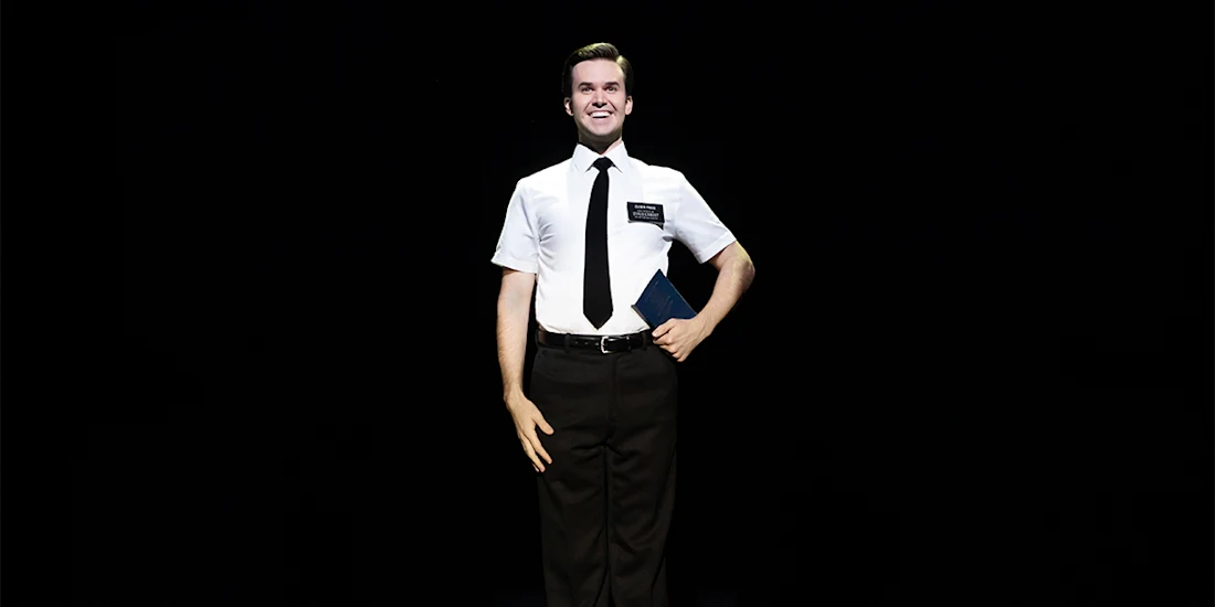 A person wearing a white short-sleeve shirt, black tie, and black pants stands and smiles on stage, holding a book against a black background.
