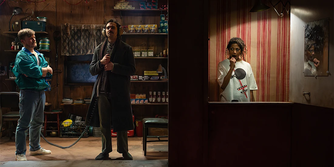 Two men stand in a cluttered shop using a corded phone, while a woman in a striped booth listens on a headset and looks into a mirror.