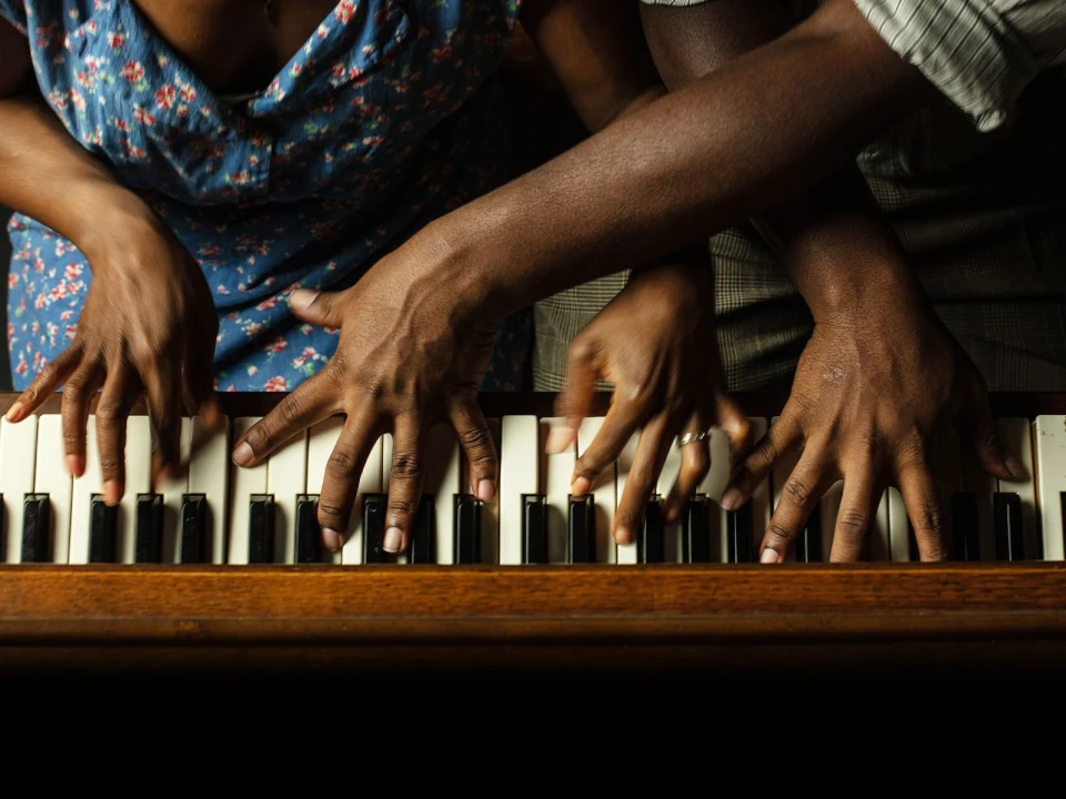 Four hands, two from each of two people, playing piano keys simultaneously. One person wears a floral dress, the other a striped shirt.