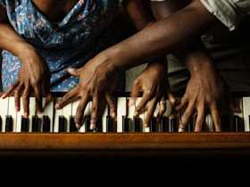 Four hands, two from each of two people, playing piano keys simultaneously. One person wears a floral dress, the other a striped shirt.