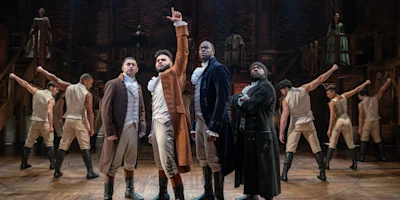 Four actors in historical costumes stand at the front of a stage, with one raising a finger, while ensemble dancers pose in the background, set against a brick backdrop.