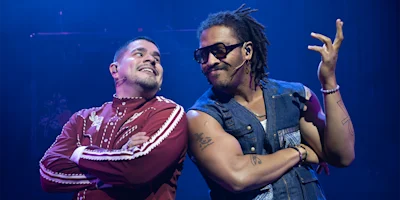 Two actors stand on stage under blue lighting, one in a red tracksuit with arms crossed, the other in a sleeveless denim vest and sunglasses, gesturing with his hand.