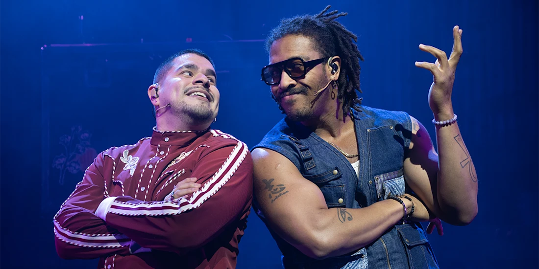 Two actors stand on stage under blue lighting, one in a red tracksuit with arms crossed, the other in a sleeveless denim vest and sunglasses, gesturing with his hand.