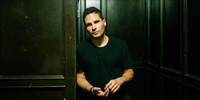 A man in a black t-shirt stands in a dimly lit room with dark wooden paneled walls, looking at the camera with a neutral expression.