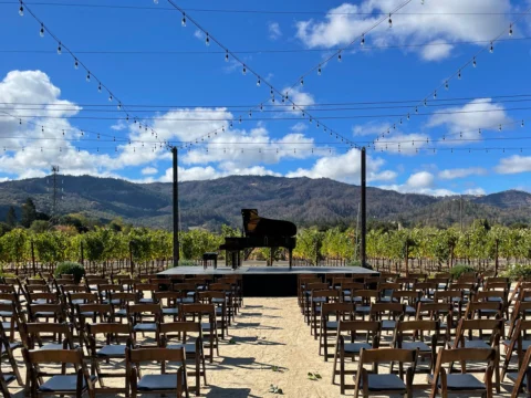 Outdoor event setup with a grand piano on a stage, surrounded by rows of empty wooden chairs. String lights are hanging above, and a mountainous landscape is visible in the background under a blue sky.