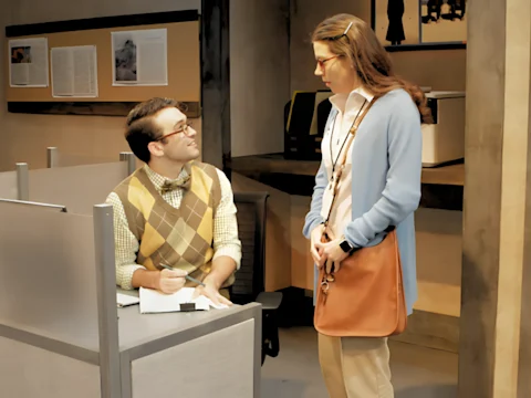 A man and a woman in an office setting. The man is sitting at a desk with a notebook, wearing glasses and a bow tie. The woman, with long hair and glasses, stands holding a bag.