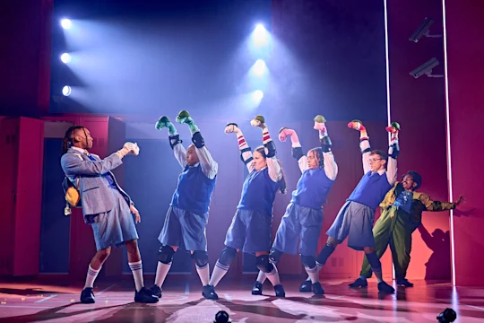 A group of performers in school uniforms and knee pads strike dynamic poses on stage under bright lights, with one person in green standing to the side.