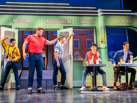 Five people are on stage dressed in mid-20th-century attire, set in a diner with signs for sundaes, sandwiches, coffee, and malts. Two are sitting at a table, and three are entering or exiting doors.