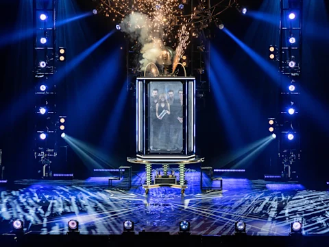 Two people stand inside a transparent water tank on a lit stage, surrounded by spotlights and smoke during a live performance.