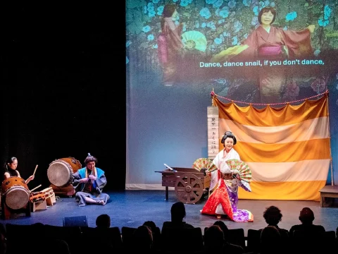 A performer in traditional attire dances on stage with colorful fans, accompanied by musicians playing drums.