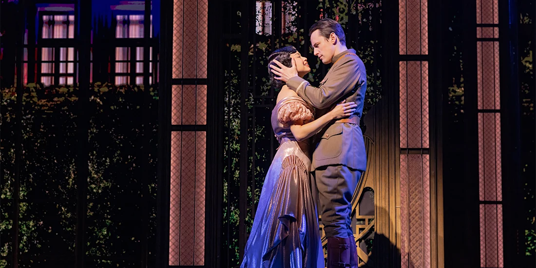 Two actors on stage in an emotional embrace, with one dressed in a formal gown and the other in a military uniform, set against a backdrop of windows and foliage.