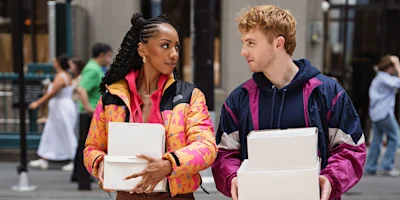 Two people walk side by side outdoors, each carrying white boxes and wearing colorful jackets, while looking at each other.