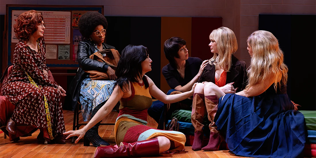 Six women sit in a circle on a gym floor, engaged in conversation. Some appear attentive while others look contemplative. Colorful posters are visible in the background.
